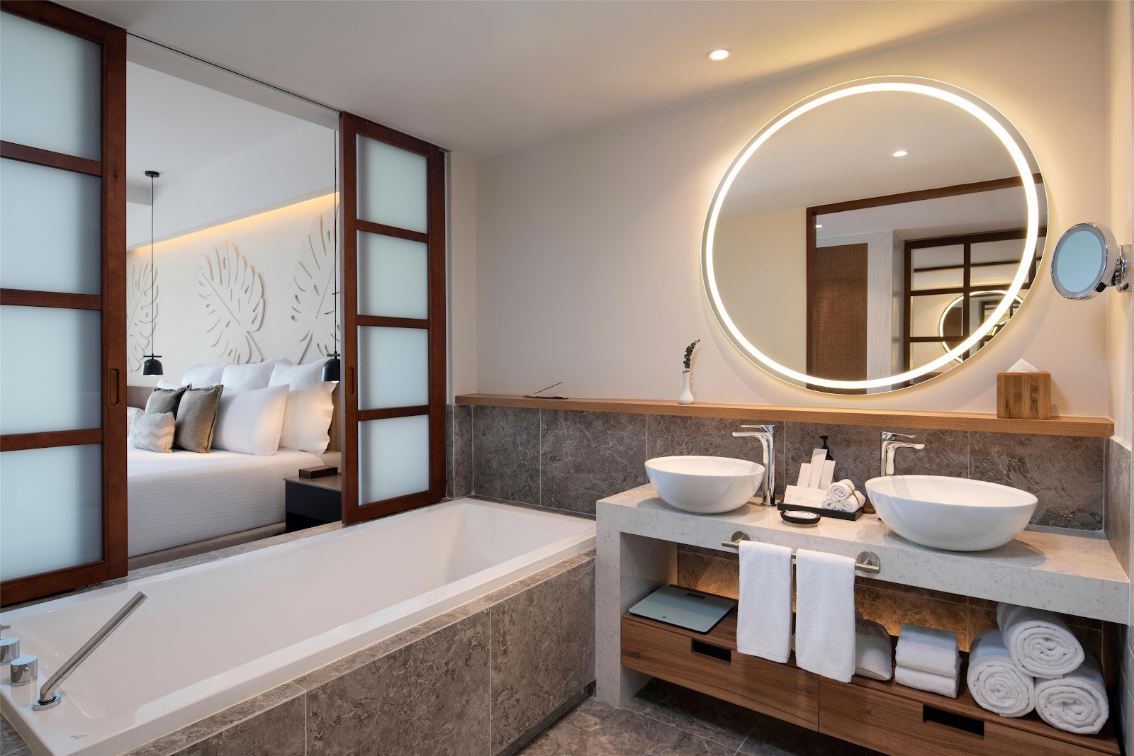 Spa bathroom with soaking tub, double vessel sinks, illuminated circular mirror, and open view to bedroom at Secrets Playa Esmeralda.