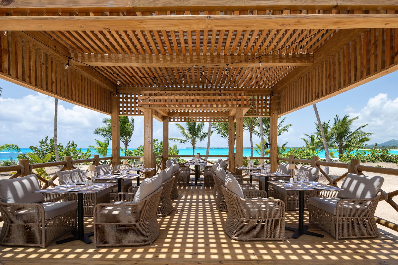 Wooden pergola dining pavilion with woven chairs overlooking turquoise ocean and palm trees at Secrets Playa Esmeralda.