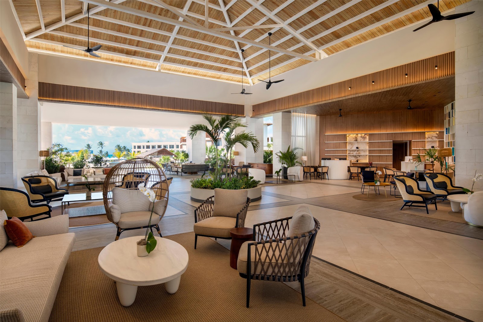 Spacious lobby at Secrets Playa Esmeralda with wooden ceiling, modern furniture, potted palms, and ocean view through open archway.
