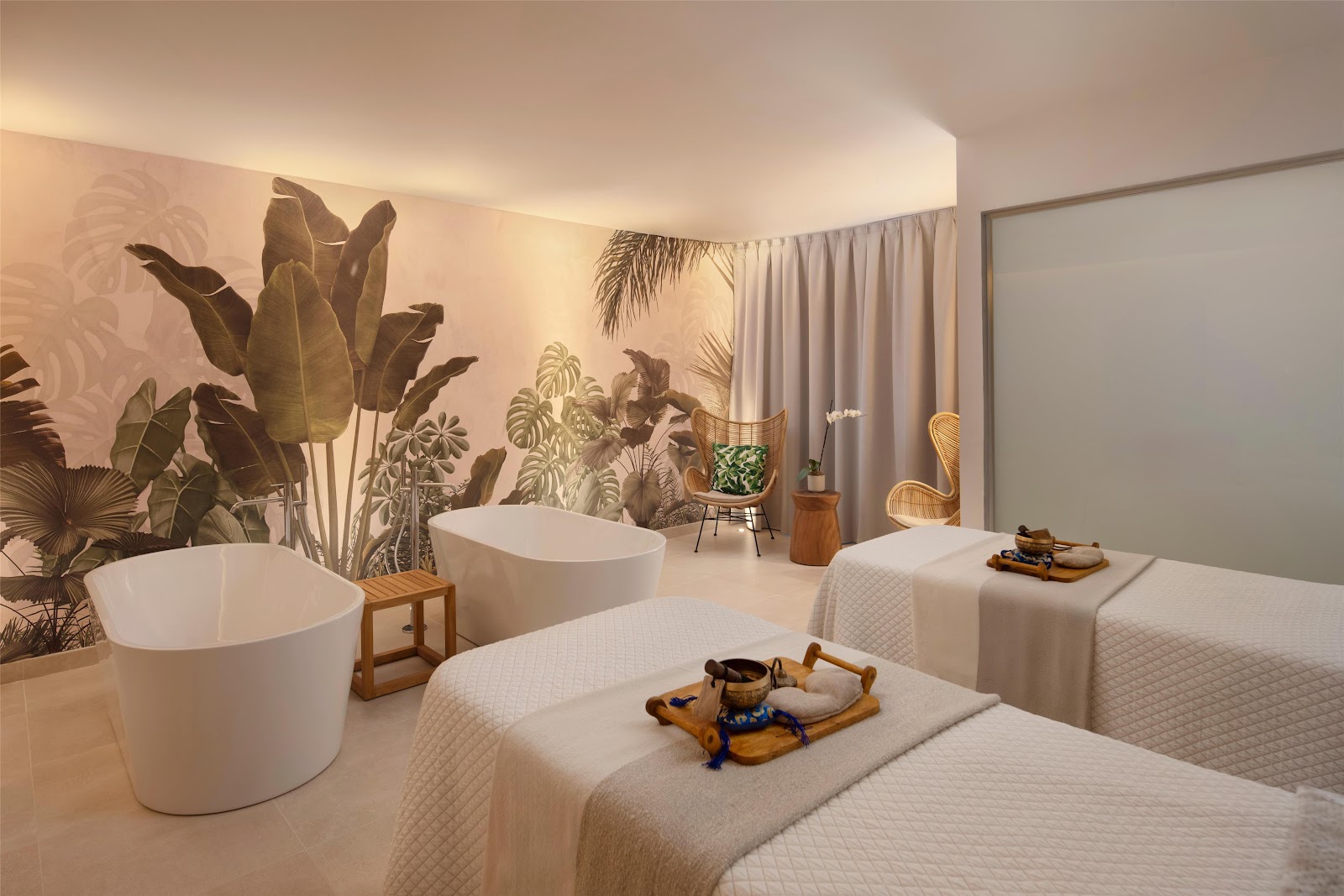 Spa treatment room at Secrets Playa Esmeralda with soaking tubs, massage beds, botanical mural, and woven chairs.