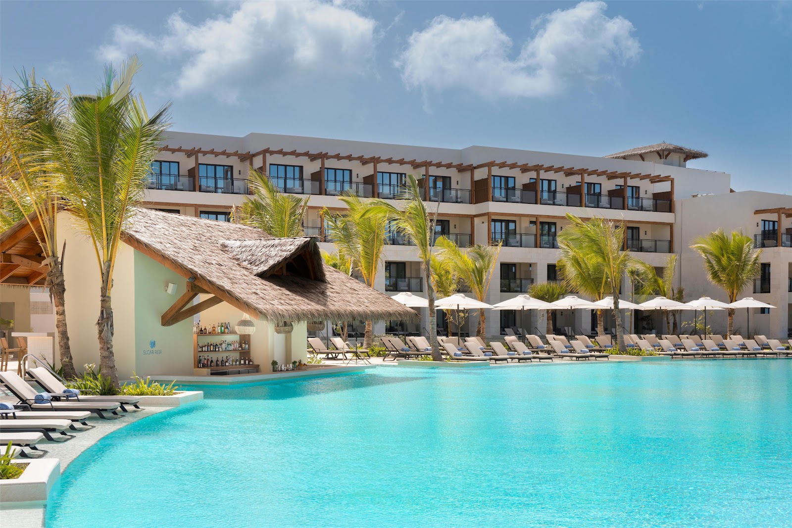 Turquoise pool with lounge chairs and thatched-roof bar at Secrets Playa Esmeralda, with multi-story resort building and palm trees beyond.