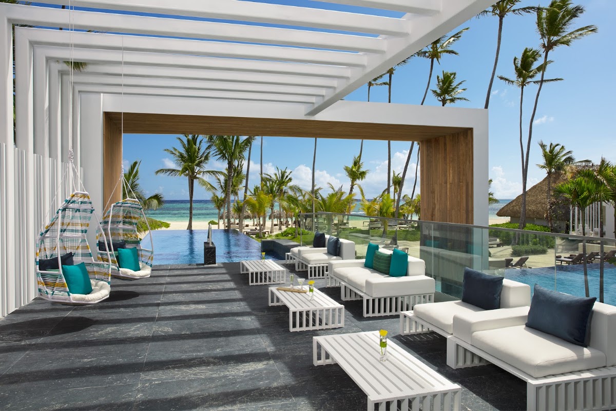 Modern lounge area with white seating, hanging chairs, and infinity pool overlooking palm trees and tropical beach at Secrets Royal Beach Punta Cana.