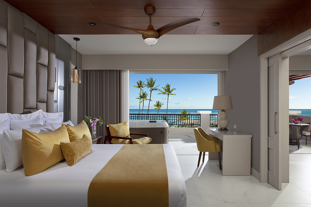 Beachfront resort room with king bed, yellow accent pillows, ocean and palm tree views through sliding glass doors, and balcony overlooking the beach.