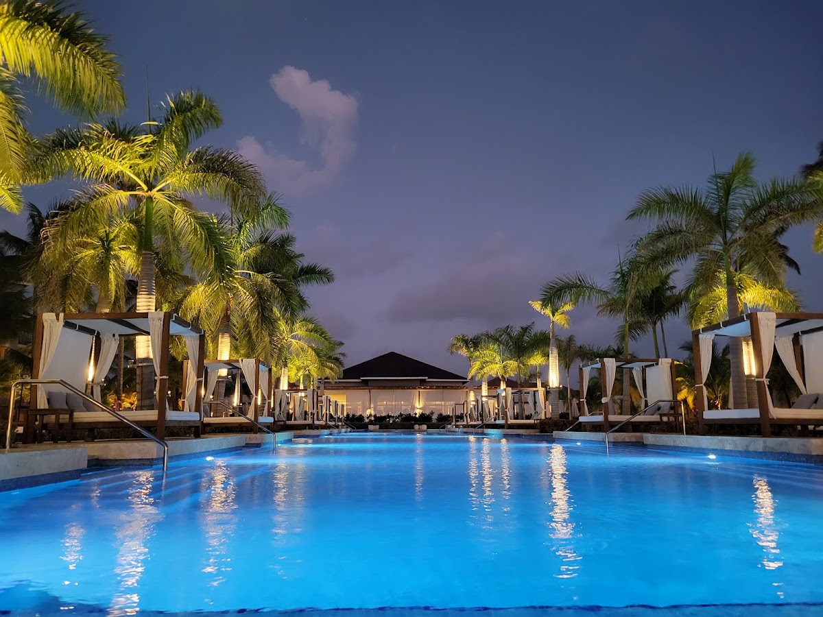 Illuminated pool at dusk with white cabanas, palm trees, and a central structure under a twilight sky at Secrets Royal Beach Punta Cana.