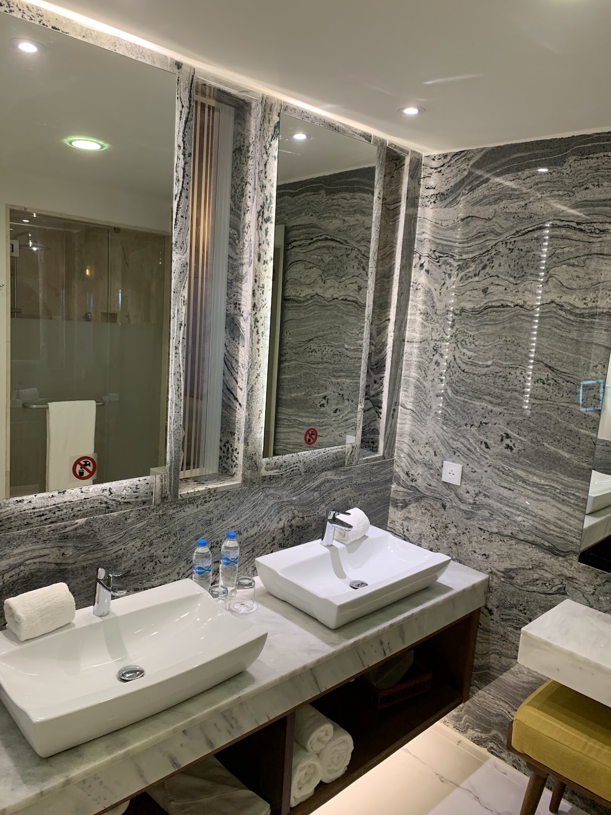 Modern bathroom with dual vessel sinks, marble countertop and walls, large illuminated mirrors, and glass shower enclosure.
