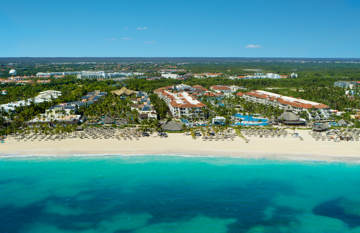 Aerial view of Secrets Royal Beach Punta Cana resort along a sandy beach with turquoise water, palm trees, and buildings inland.