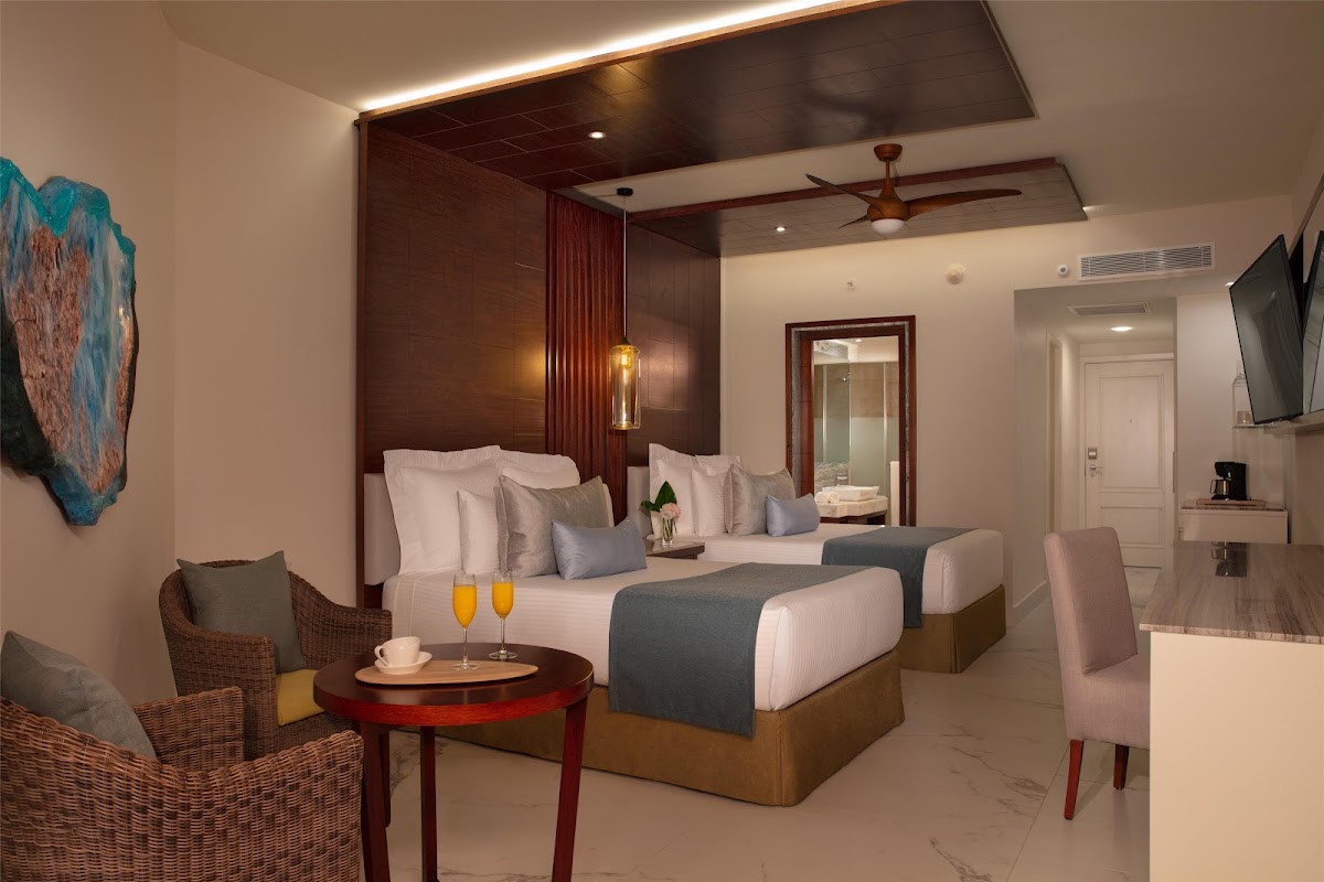 Spacious resort room with two queen beds, dark wood headboard, wicker seating, and champagne glasses on a side table.