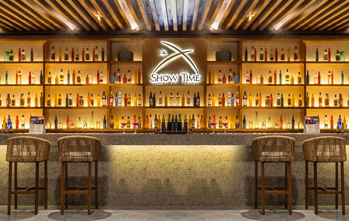 Bar counter with woven stools and illuminated shelves displaying liquor bottles above a neon "Show Time" sign.