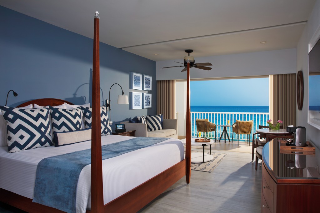 Bedroom with four-poster bed, blue accent wall, and ocean view through open doors to a balcony with two chairs.