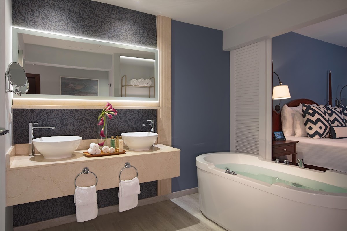 Bathroom with dual white vessel sinks on a light wood vanity, illuminated mirror, dark patterned tile backsplash, soaking tub with green water, and adjoining bedroom with navy accent wall.