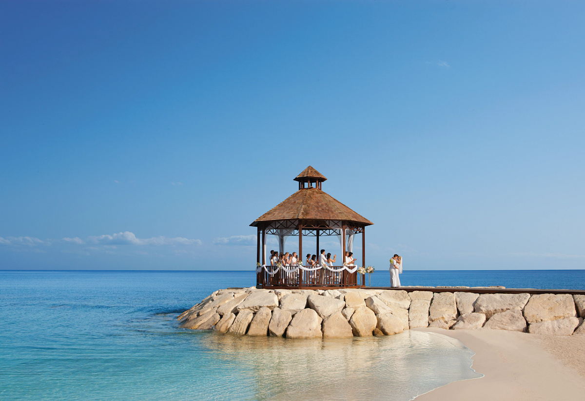 A wooden gazebo with a peaked roof sits on a rocky jetty in clear turquoise water, with guests gathering inside and on the beach below under blue sky.