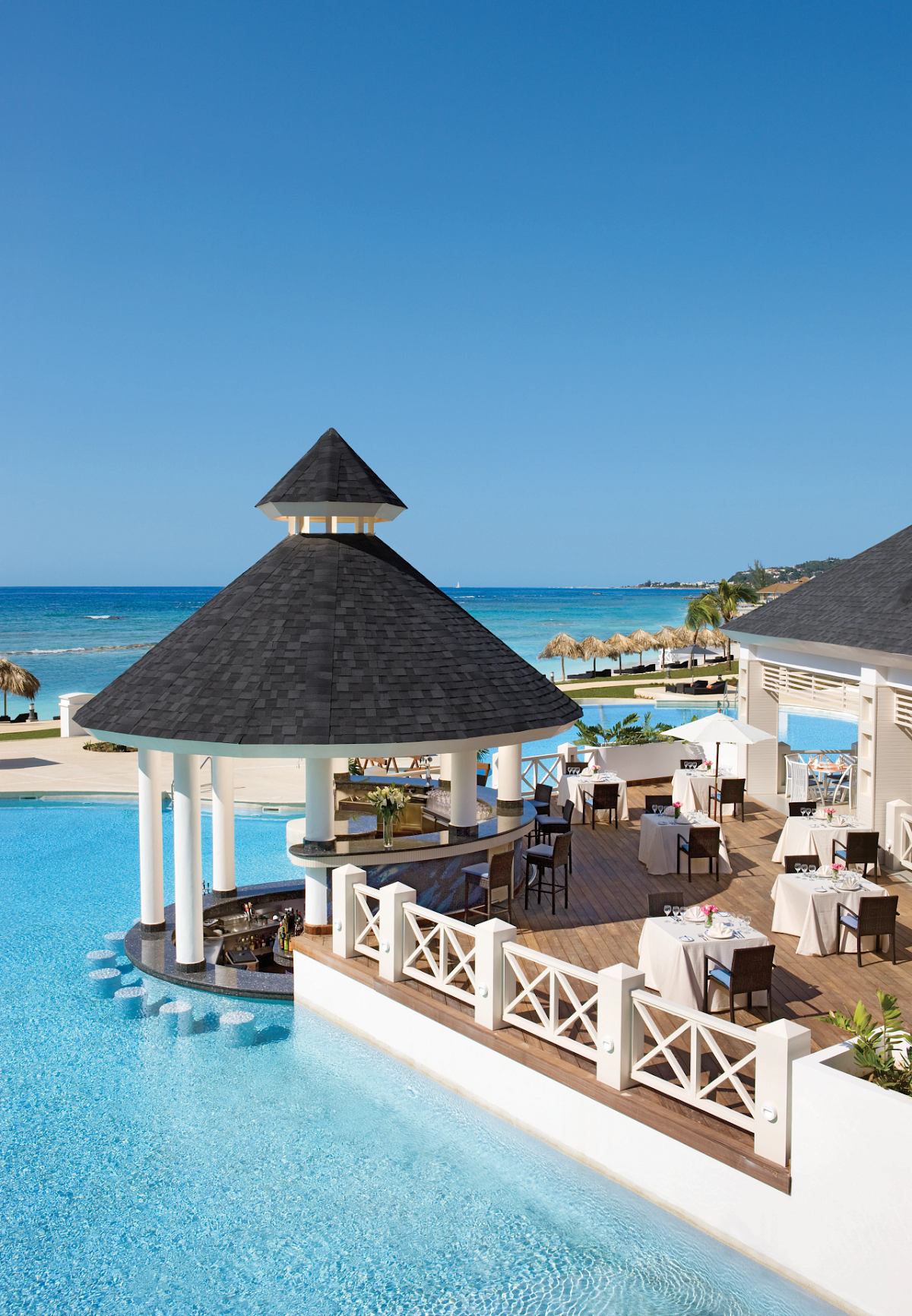 Beachfront pool bar with a black conical roof and white columns overlooks the turquoise ocean at Secrets St. James Montego Bay.