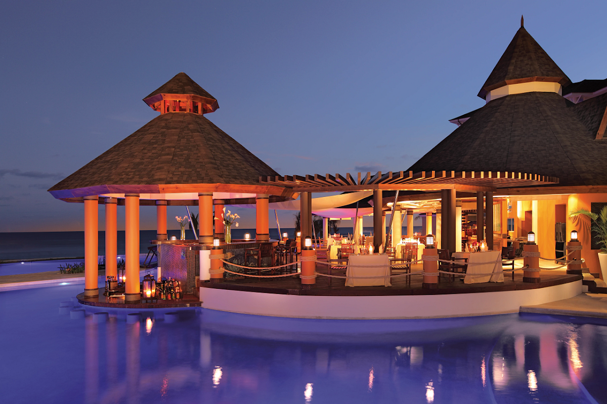 Illuminated oceanfront pavilions with conical roofs and orange columns overlook a glowing pool at dusk, with the ocean beyond.