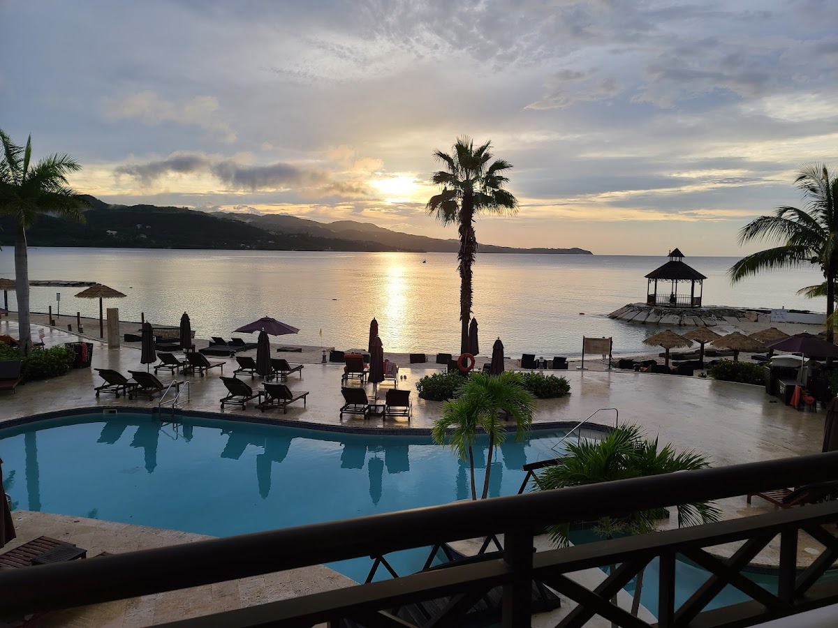 Resort pool overlooking Montego Bay at sunset with loungers, umbrellas, and a wooden gazebo on the beach beyond.
