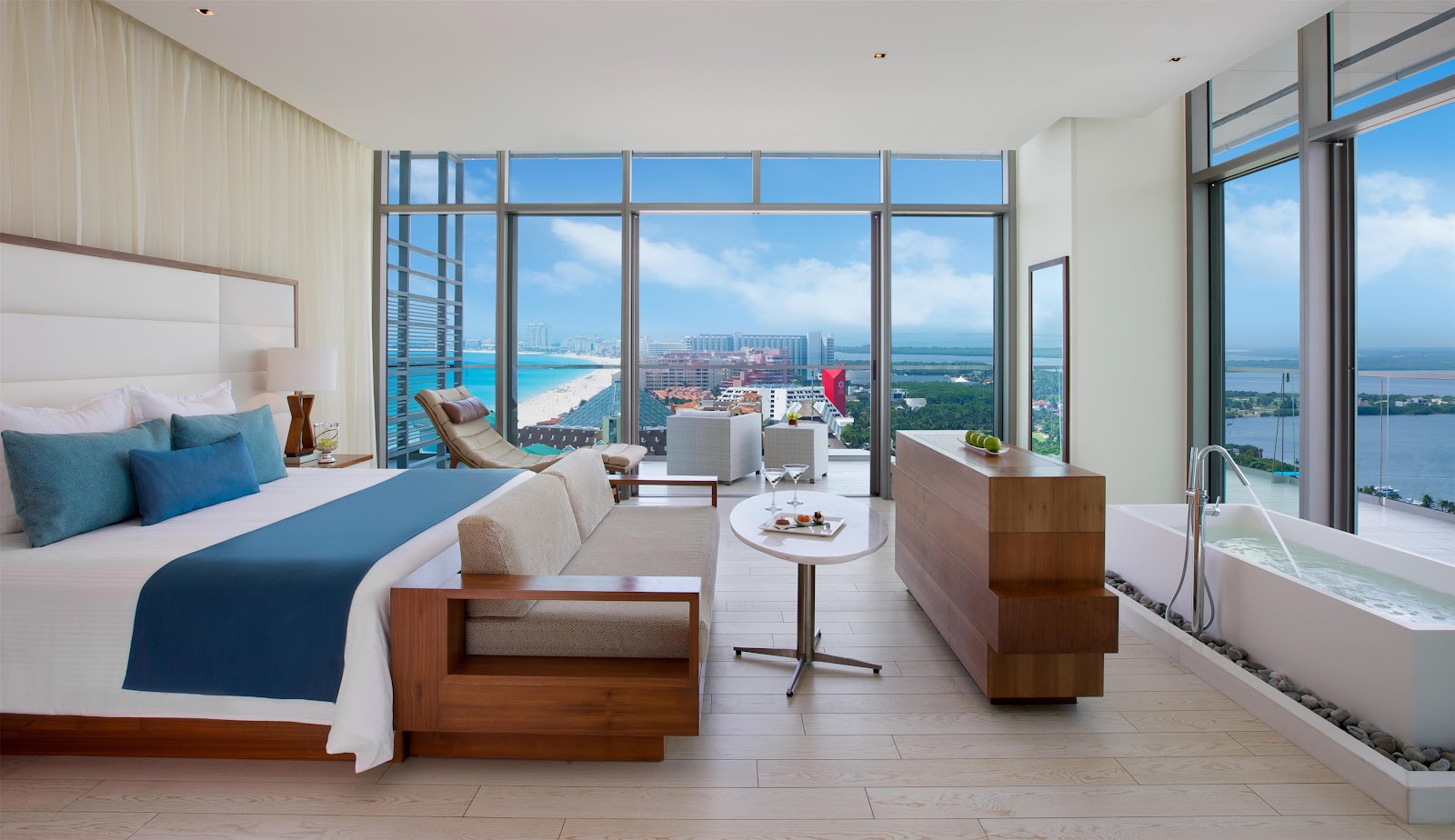 High-rise resort suite with king bed, lounge seating, and freestanding soaker tub overlooking Cancun's turquoise Caribbean waters and beach from floor-to-ceiling windows.