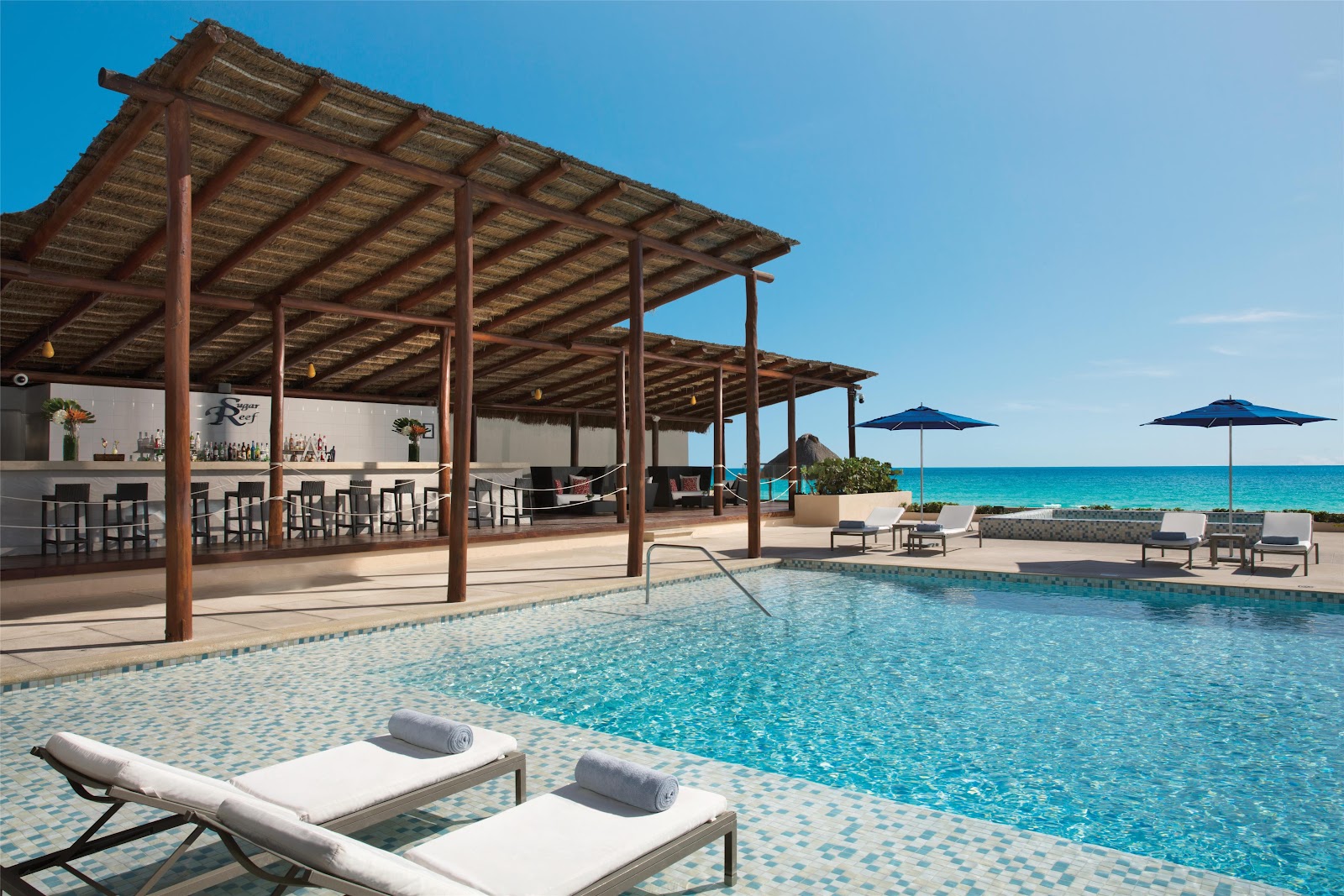 Pool area with wooden pergola, bar, lounge chairs, and turquoise ocean view under clear sky at Secrets The Vine Cancun.