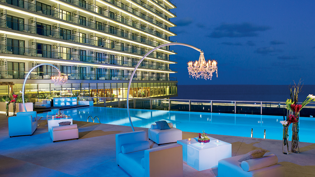 Illuminated poolside lounge at dusk with glowing chandeliers, modern blue furniture, and a multi-story resort building overlooking the ocean.