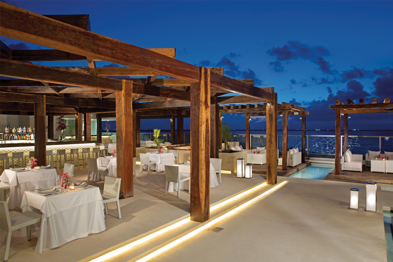 Rooftop dining area at Secrets The Vine Cancun with wooden pergolas, white-clothed tables, and ocean views at dusk.
