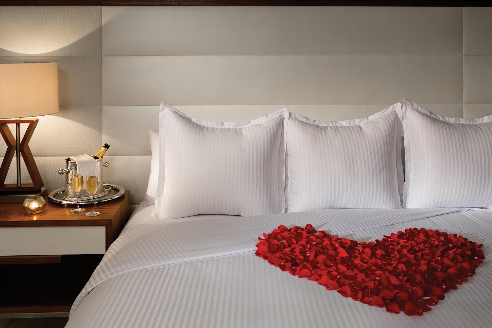 White striped bed with red rose petals arranged in a heart shape, champagne bottle and glasses on nightstand with lamp.