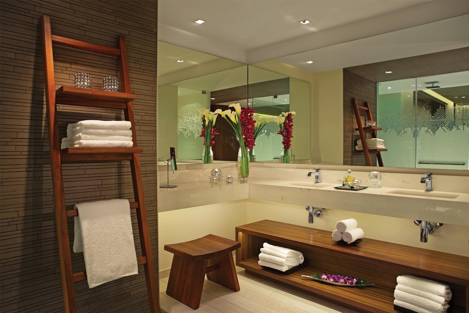 Spa bathroom with dual vanity, large mirror, wooden accents, white towels on ladder shelf, red flowers in vases, and cream-colored tile flooring.
