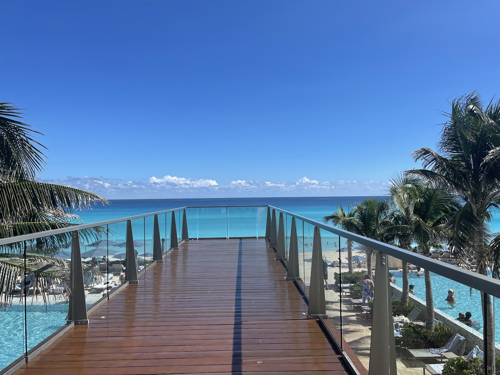 Wooden boardwalk with glass railings extending toward the ocean, flanked by palm trees and resort pools below under clear blue sky.