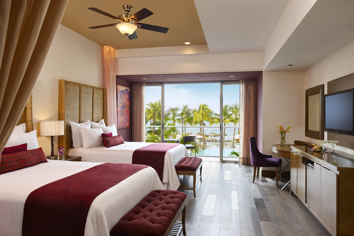 Two wooden beds with white linens and magenta pillows in a resort room with slatted wood walls and wine bottles on the nightstand.