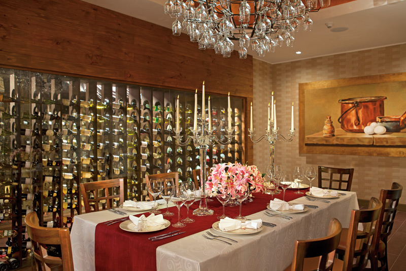 Formal dining room at Secrets Vallarta Bay with a set table featuring red and white linens, pink floral centerpiece, crystal chandelier, and wine display wall.