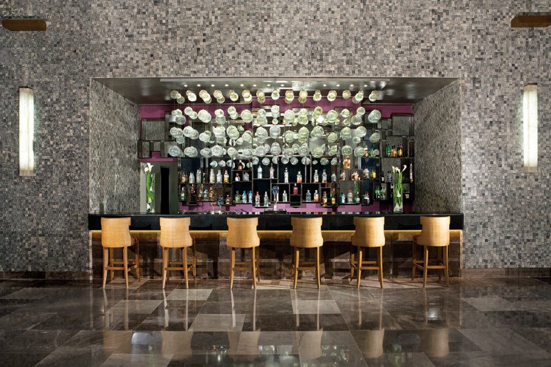 Modern bar with gray mosaic tile walls, wooden stools, extensive liquor selection, and hanging white light fixtures overhead.