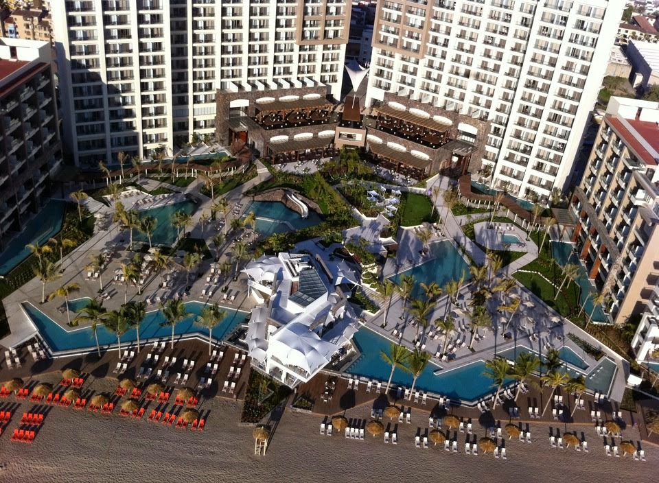Aerial view of Secrets Vallarta Bay resort with multiple pools, loungers, palm trees, and high-rise buildings surrounding the beachfront property.