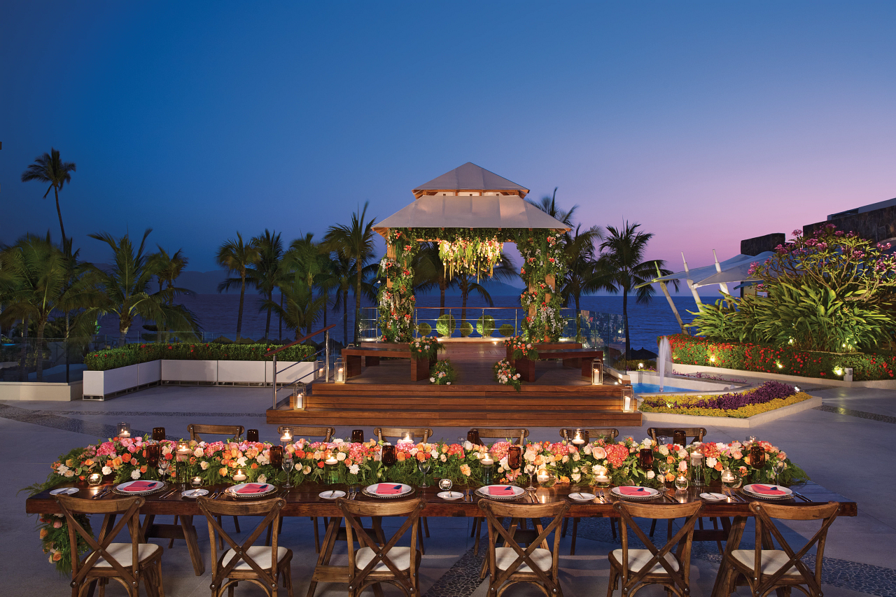 Dining tables with tan linens and fresh flowers overlook a garden and ocean view at Secrets Vallarta Bay Puerto Vallarta.