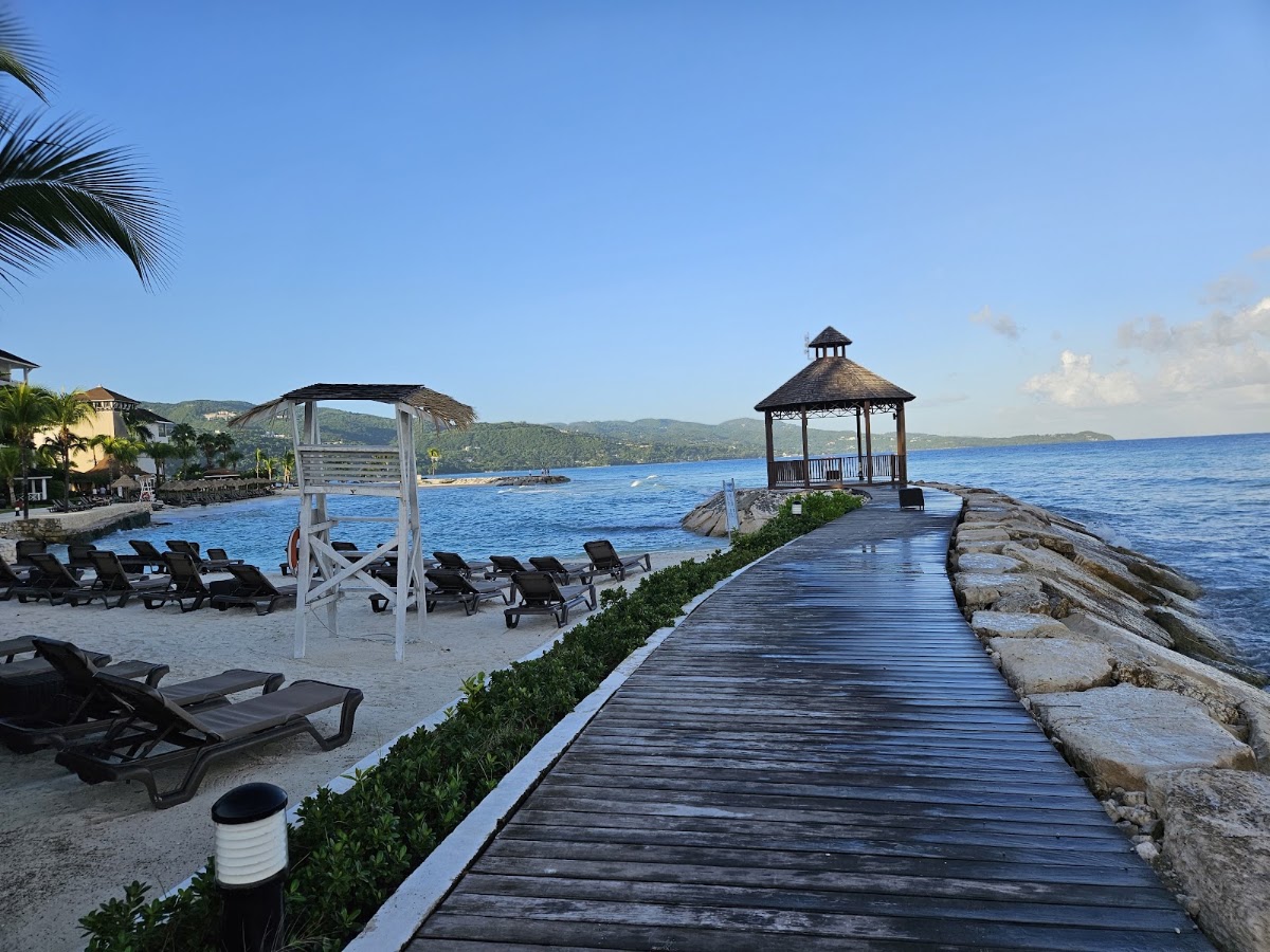 Wooden boardwalk leading to a gazebo on a rocky point, with lounge chairs and beach cabanas along the sandy shore at Secrets Wild Orchid Montego Bay.