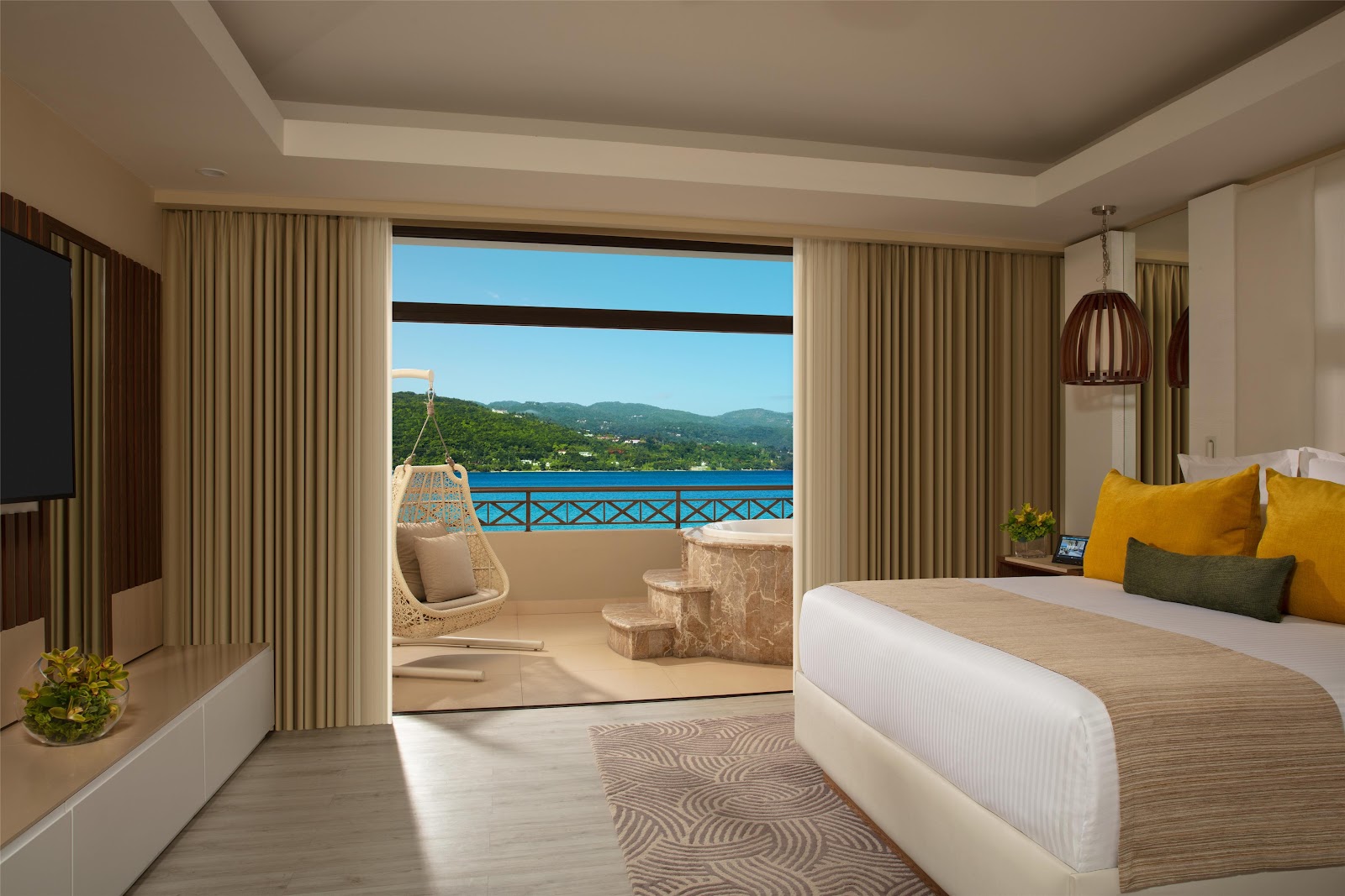 Resort bedroom with king bed, open balcony door revealing ocean view, hanging chair, and forested coastline beyond.