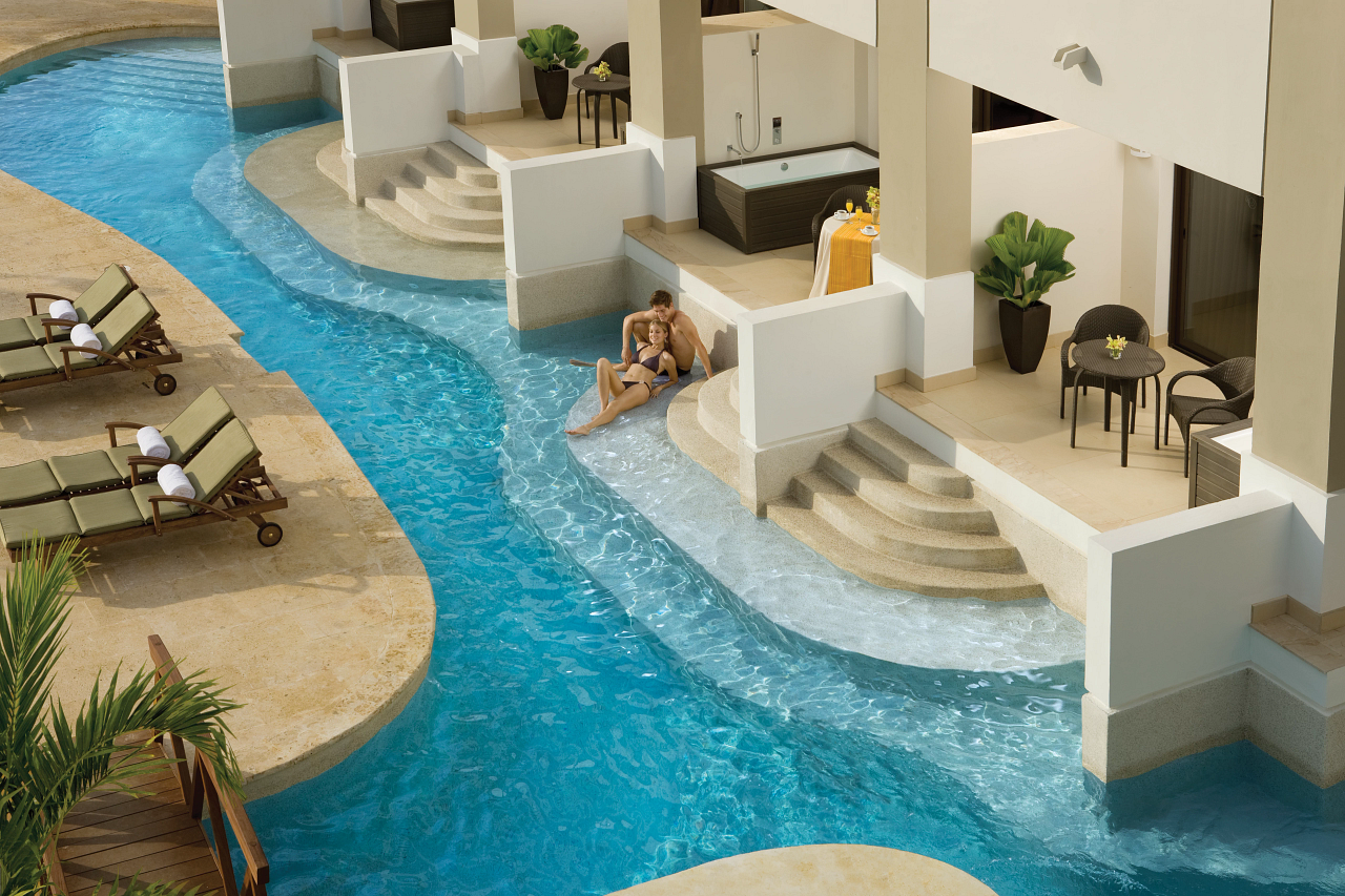 Winding lazy river flows past white resort buildings with lounge chairs and a guest wading in shallow water.