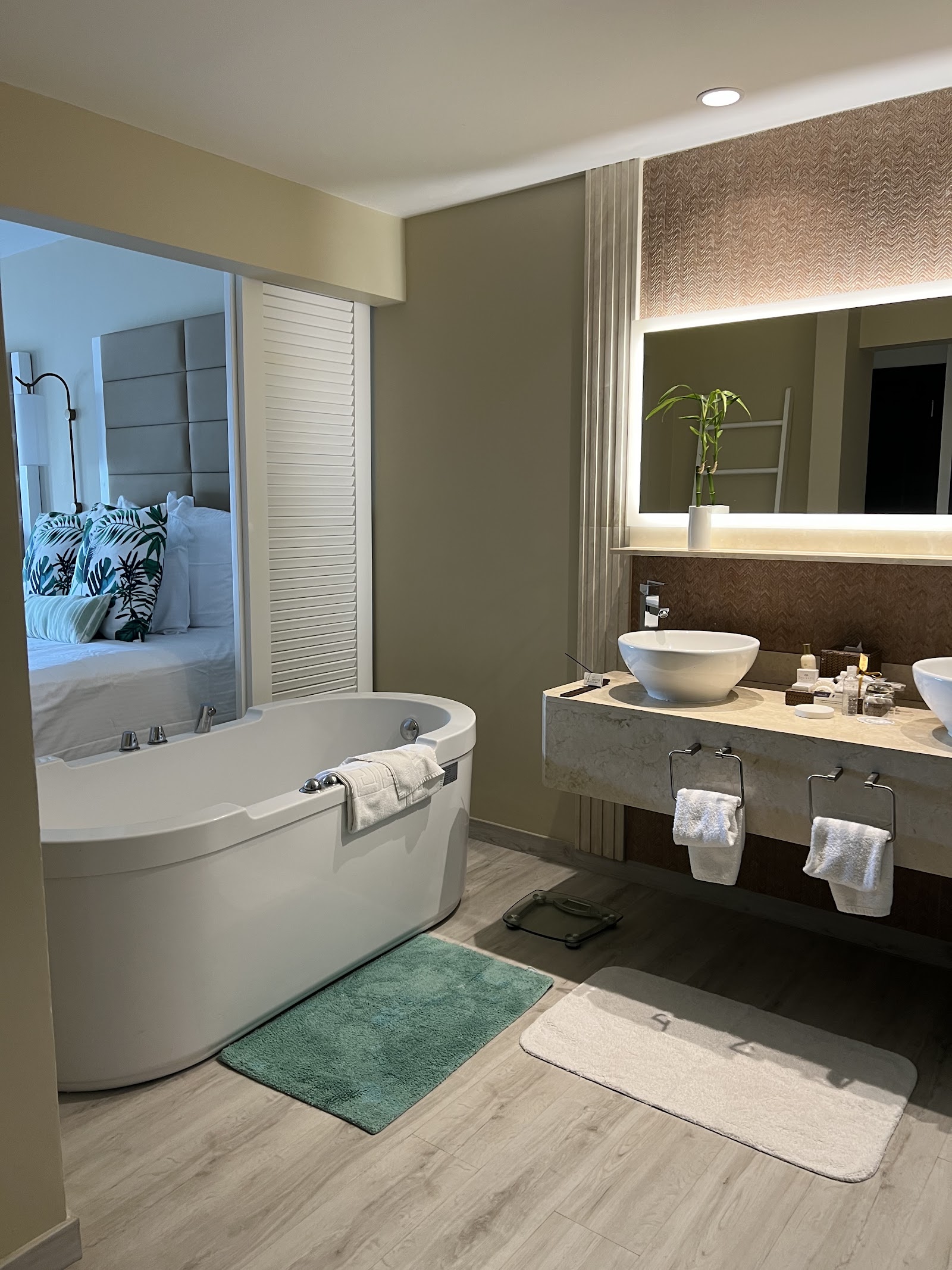 Modern resort bathroom with curved soaking tub, vessel sink, illuminated mirror, and open view into bedroom with patterned pillows.