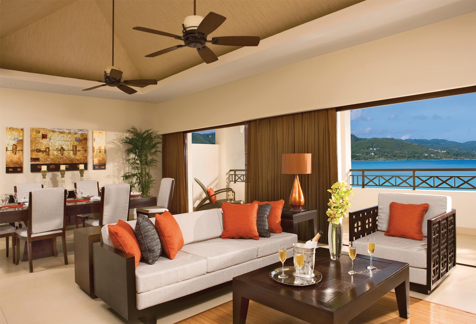 Spacious suite with cream sofa, orange accent pillows, and ocean view balcony overlooking blue Caribbean waters and forested coastline.
