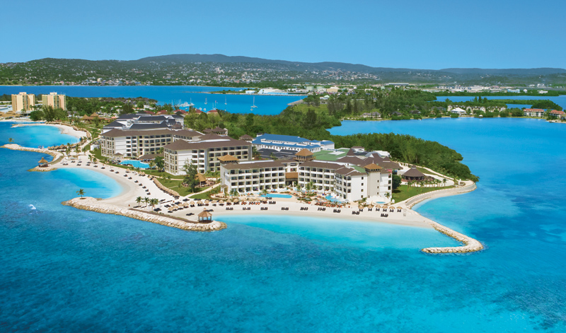 Aerial view of Secrets Wild Orchid Montego Bay resort on a peninsula surrounded by turquoise water, with sandy beach and mountainous coastline beyond.
