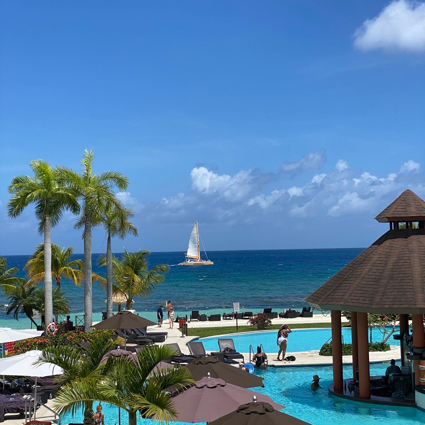 Resort pool with swim-up bar and wooden gazebo, palm trees, and sailboat on turquoise ocean under blue sky.