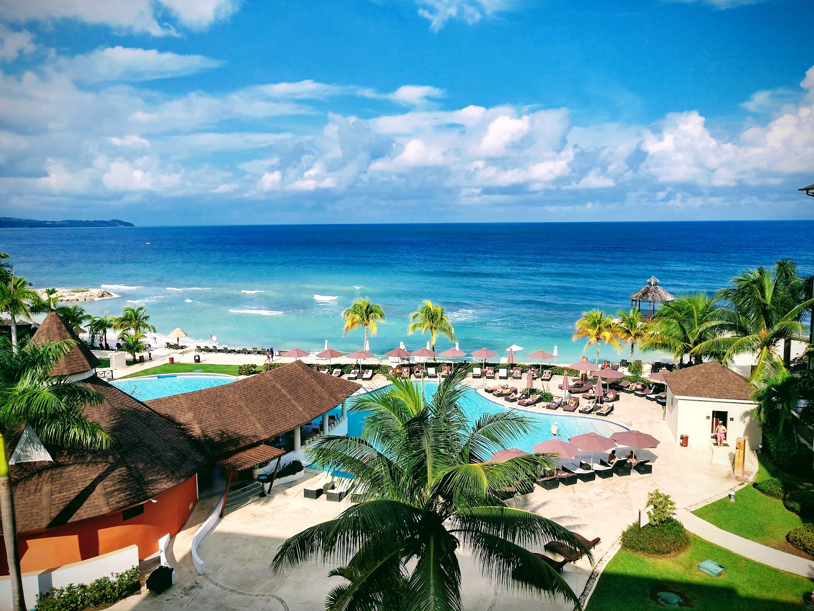 Secrets Wild Orchid Montego Bay overlooks a turquoise Caribbean beach with multiple pools, lounge chairs, and palm trees under blue sky with white clouds.
