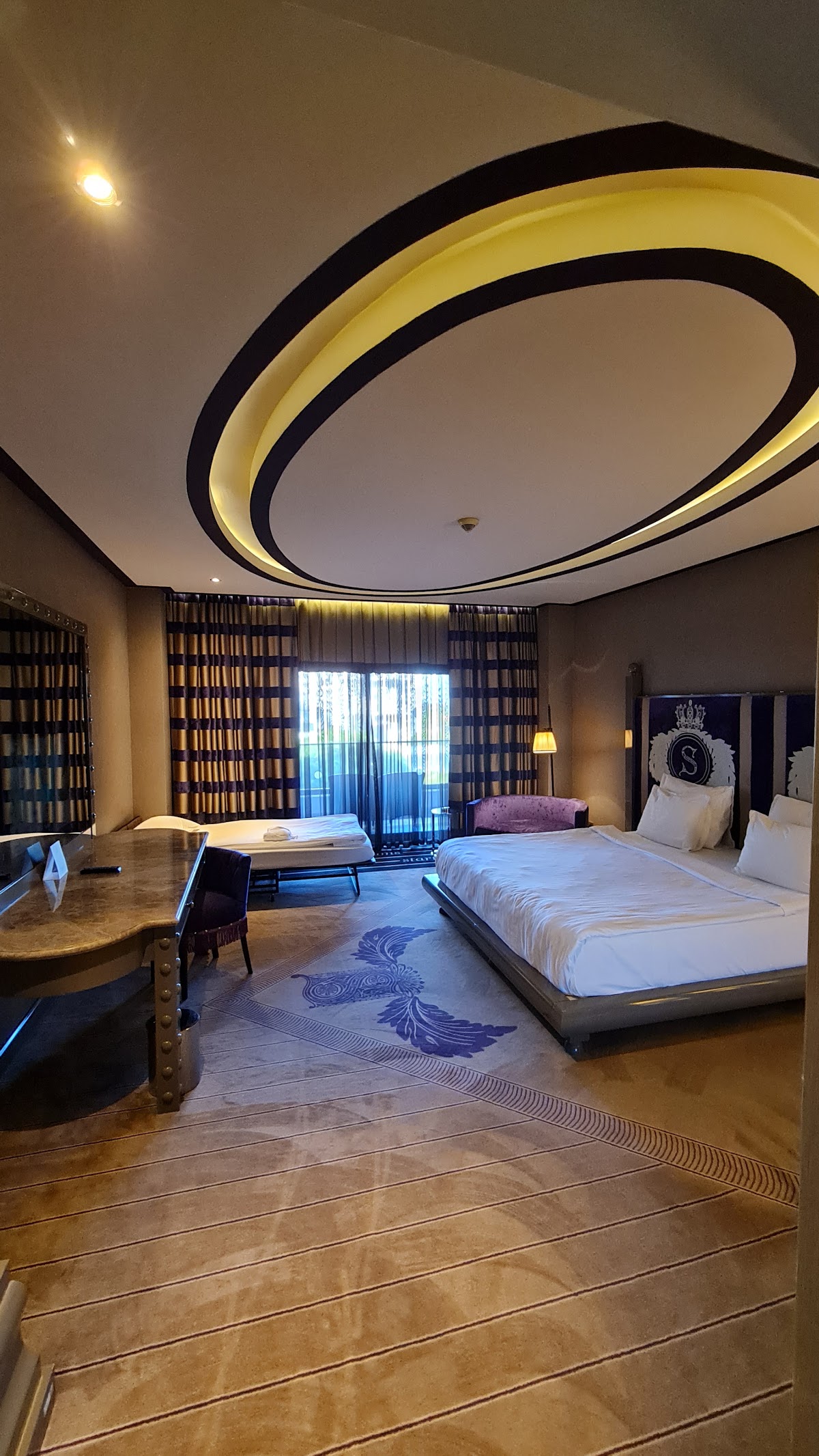 Spacious resort suite with a curved yellow and black ceiling feature, queen bed with white linens, desk area, and balcony doors with curtains.