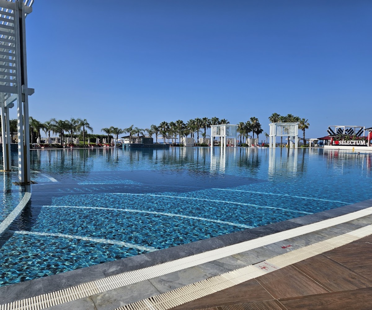 Large lagoon-style pool at Selectum Luxury Resort Belek with palm trees, white buildings, and clear blue sky reflecting on calm water.