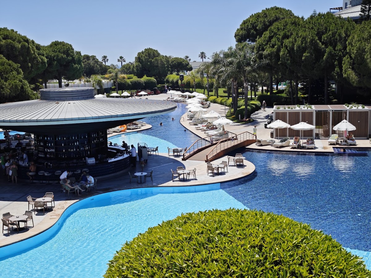 Curved pools with a swim-up bar, lounge chairs, white umbrellas, and mature trees surrounding the Selectum Luxury Resort Belek.