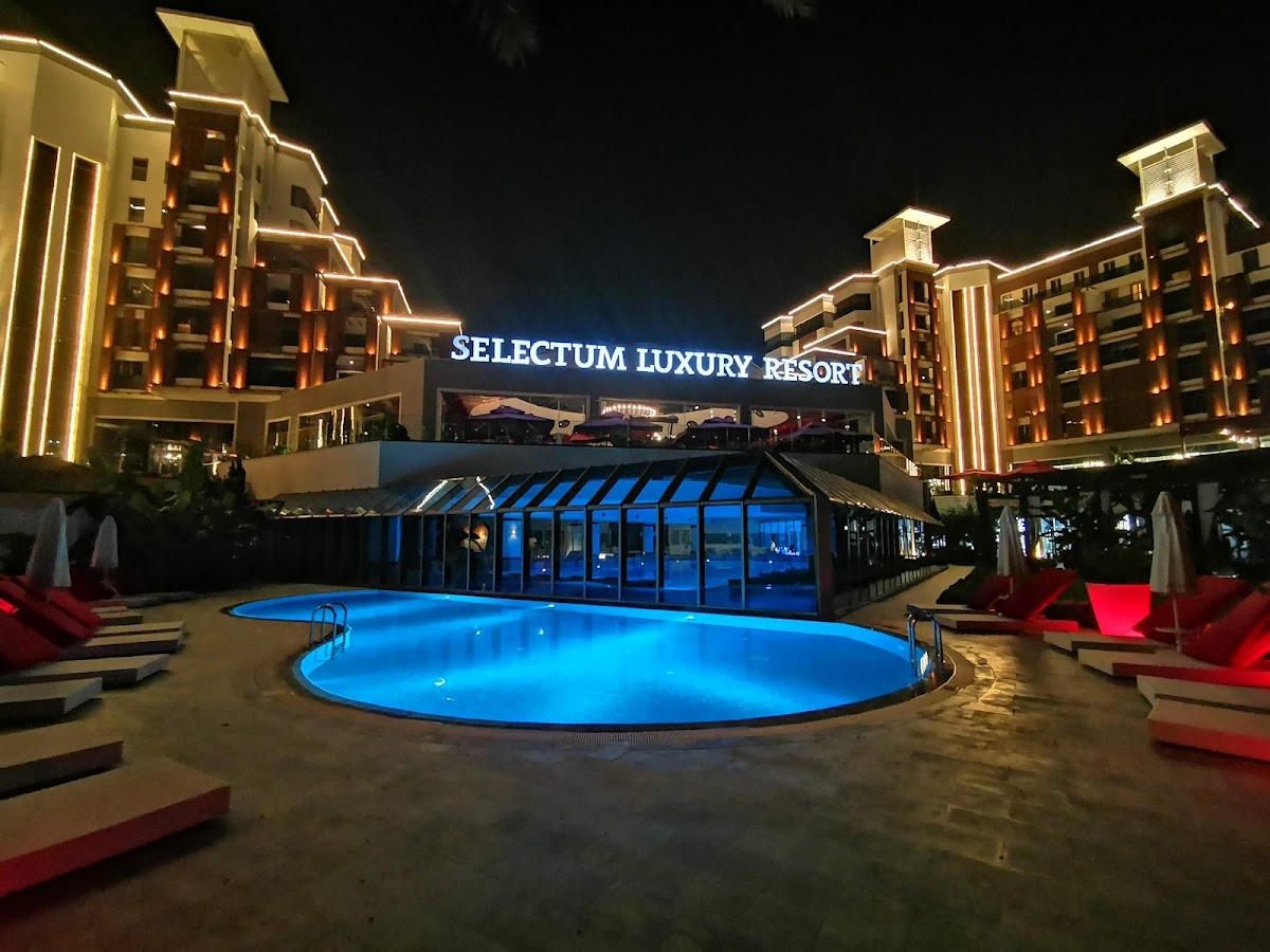 Selectum Luxury Resort's illuminated pool glows blue at night, surrounded by red lounge chairs and flanked by lit hotel buildings.