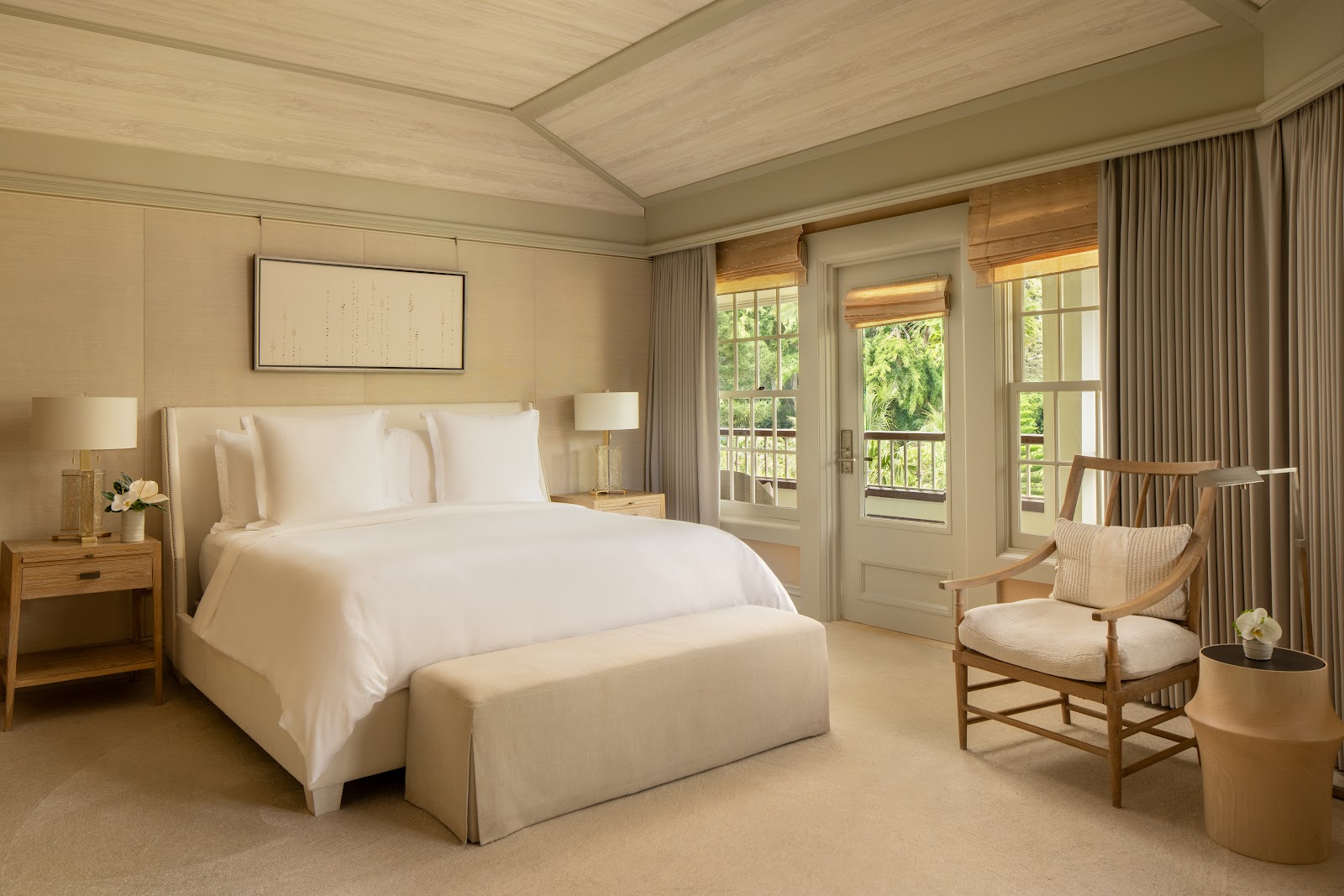 Bright bedroom with two beds, white bedding, desk with lamp, lounge chair, and floor-to-ceiling windows overlooking tropical vegetation and a wooden deck.