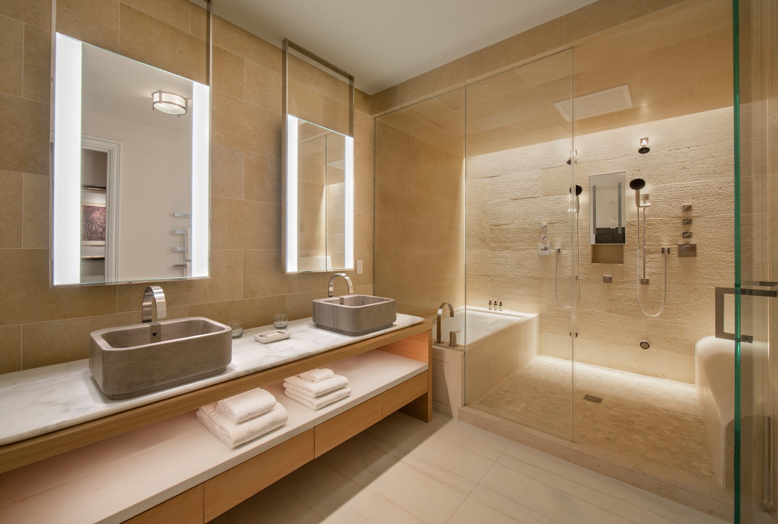 Spa bathroom with dual vessel sinks, lighted mirrors, marble countertop with folded towels, and large glass-enclosed shower with stone accents.
