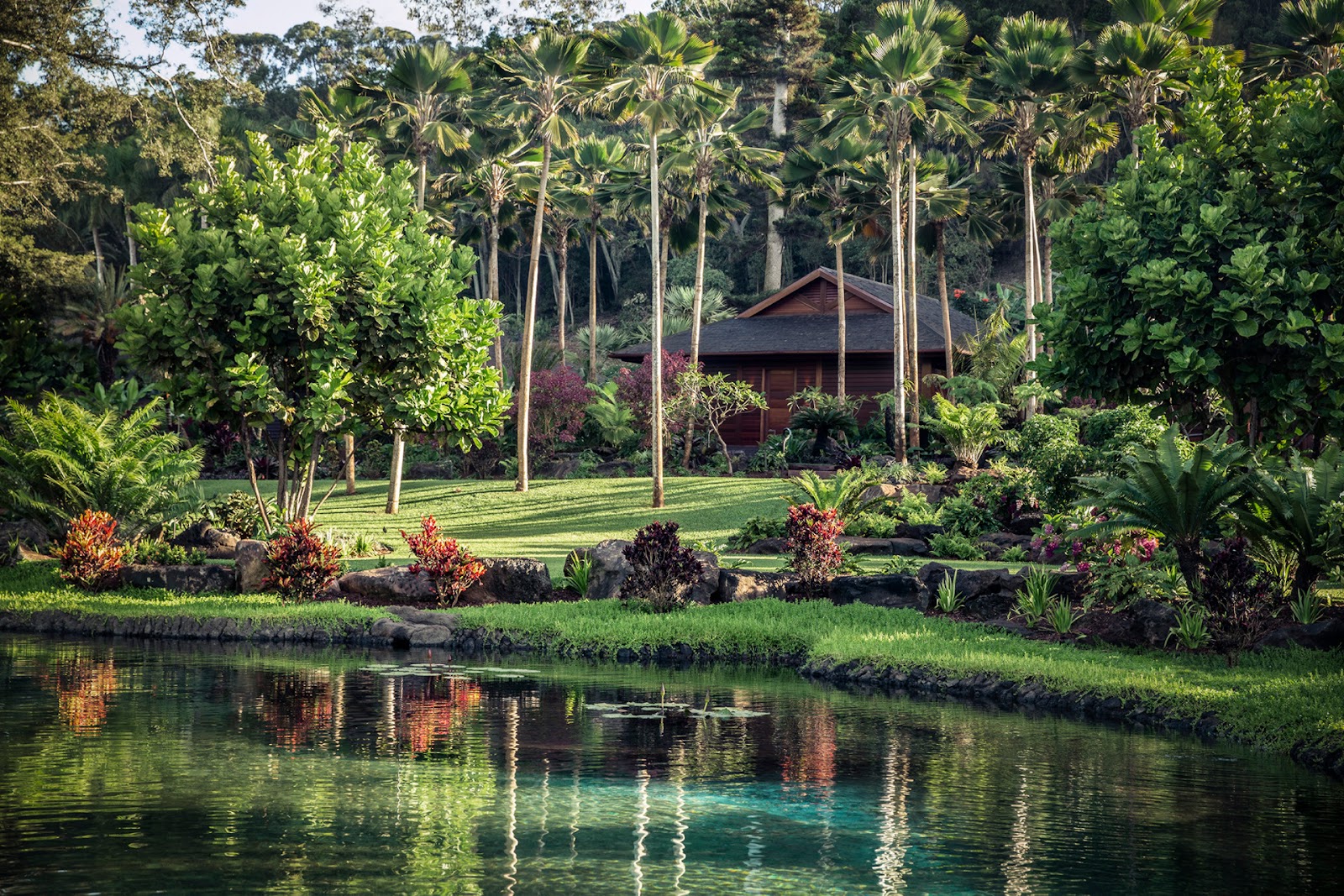 Open wooden pavilion with two cream lounge chairs and an ottoman facing tropical gardens through glass doors and windows.
