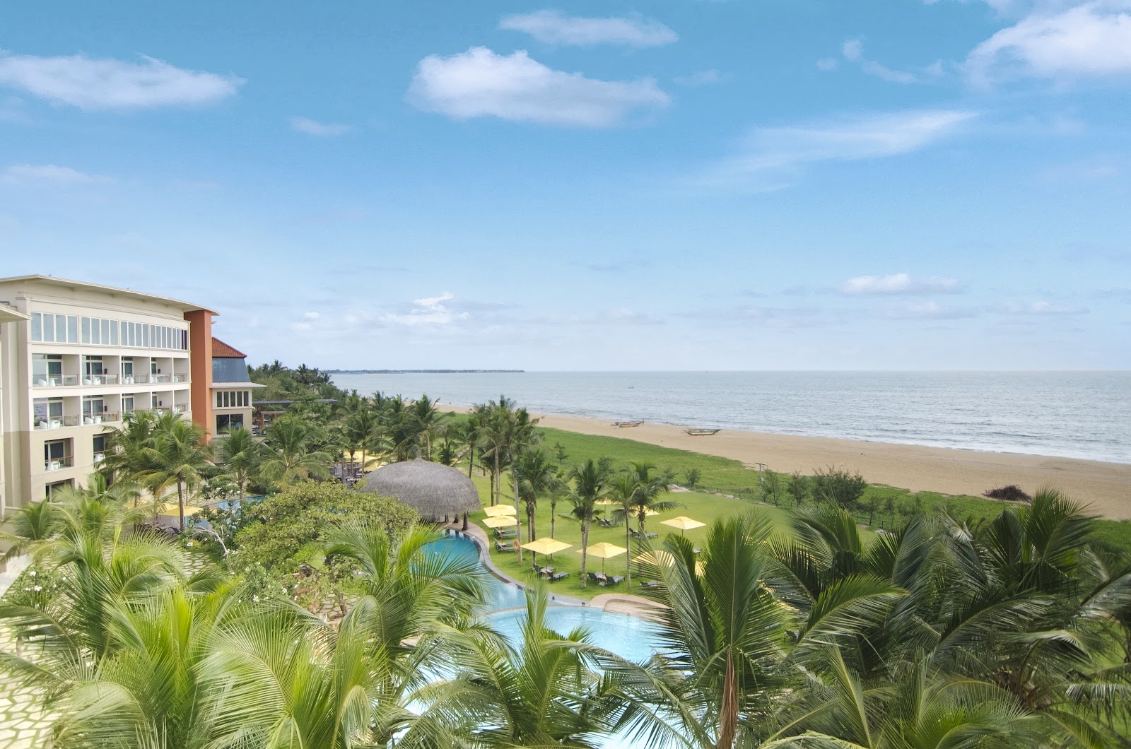 Beachfront resort with multi-story white building, palm trees, pool with thatched umbrella, and sandy beach stretching to calm ocean water under blue sky.