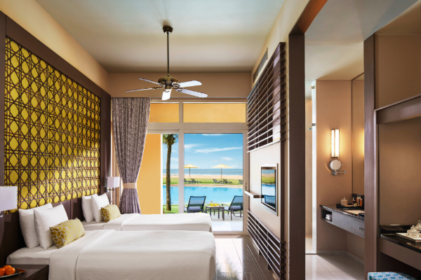 Modern resort bedroom with two beds, yellow patterned wall, and ocean view through glass doors leading to a beachfront balcony.
