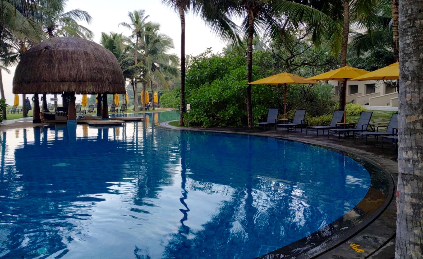 Curved swimming pool with thatched-roof bar pavilion, yellow umbrellas, lounge chairs, and palm trees at Sentido Heritance Negombo.