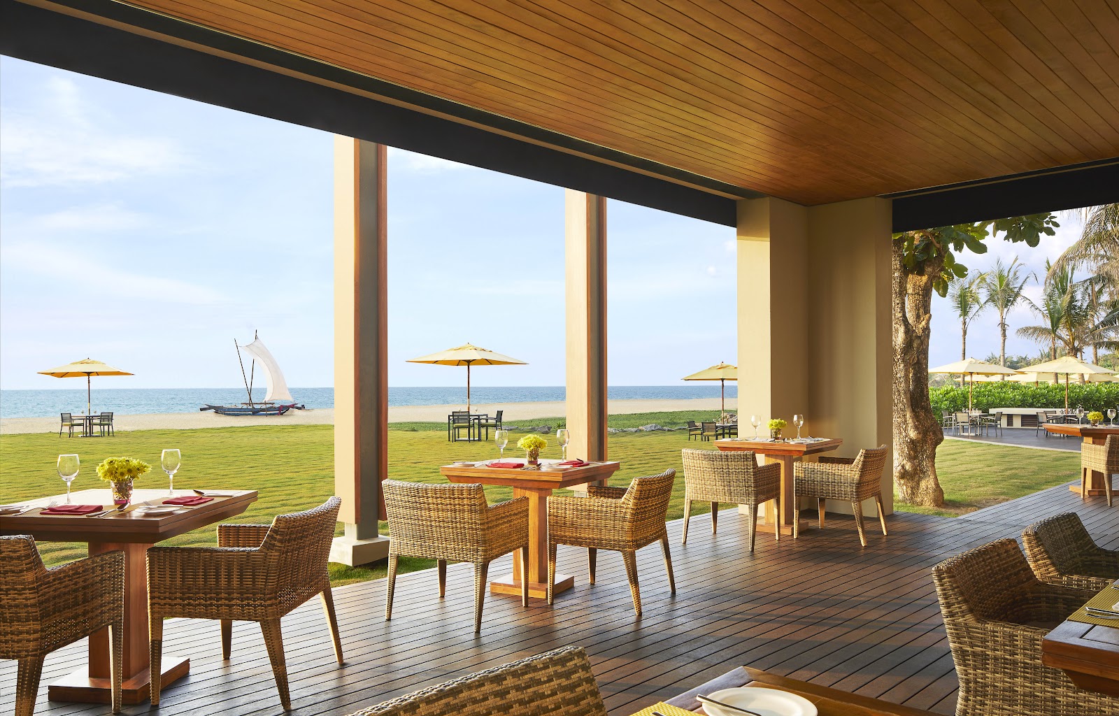 Covered beachfront dining pavilion with woven chairs, wooden tables, and ocean views, with sailboat and yellow umbrellas visible on the grass and beach beyond.