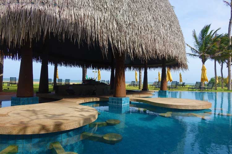 Curved pool platforms under a large thatched-roof structure at Sentido Heritance Negombo, with palm trees and ocean view beyond.